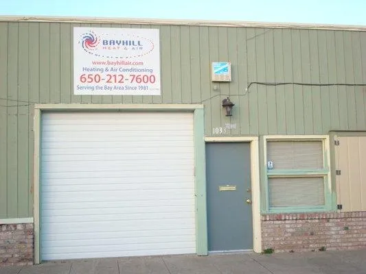 Front view of Bayhill Heat & Air building with a sign displaying contact info, garage door, and entrance. The exterior has green paneling and bricks.