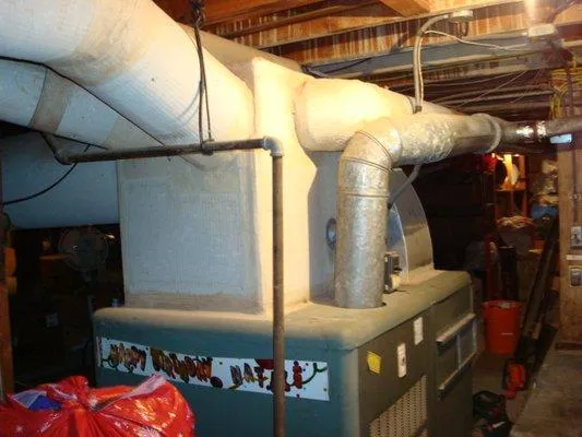 A large furnace with insulated and metal ductwork in a basement surrounded by various stored items.