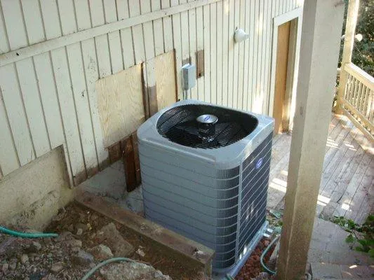 An air conditioning unit is installed outside a wooden house, next to a stairway. The unit is situated on a small concrete slab amidst some garden hoses and gravel.