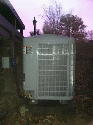  Outdoor air conditioning unit installed next to a building, surrounded by fallen leaves and trees in the background.