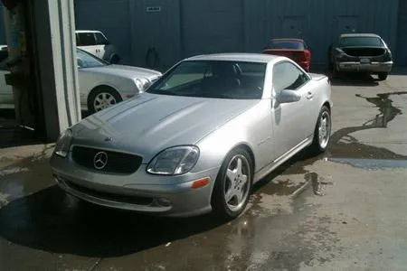 Silver car parked on a wet surface near a building, with other vehicles visible in the background.