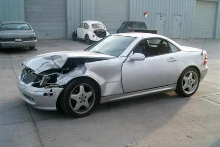  A silver car with front-end damage is parked near a building. Two other cars are visible in the background.