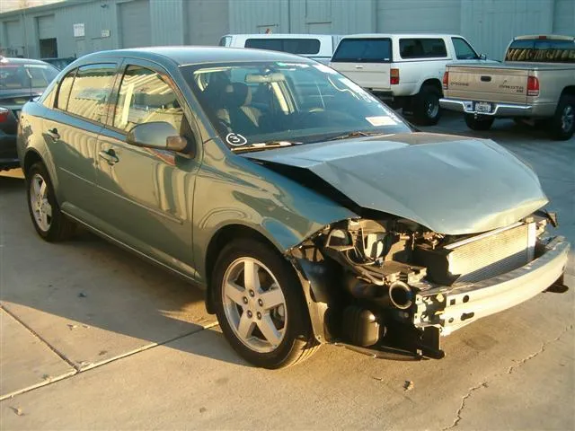 A green sedan with visible front-end damage parked in a lot with other vehicles. The car's bumper and headlight area are exposed, highlighting repair needs.