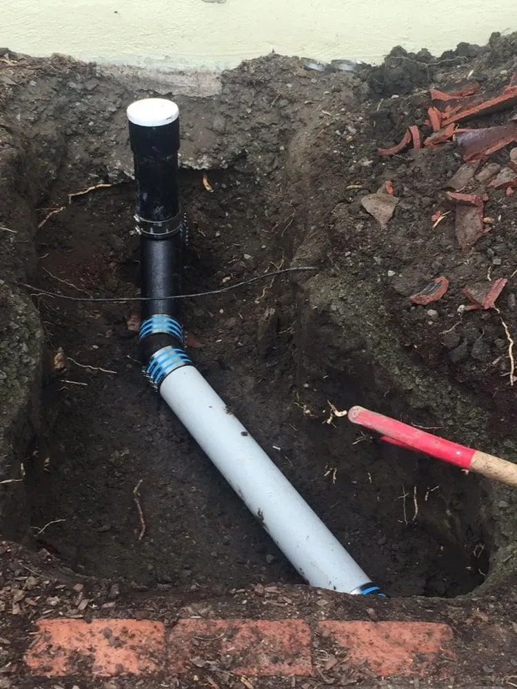 A newly installed black and white pipe in a dug trench, surrounded by soil and bricks, with a red-handled tool nearby.