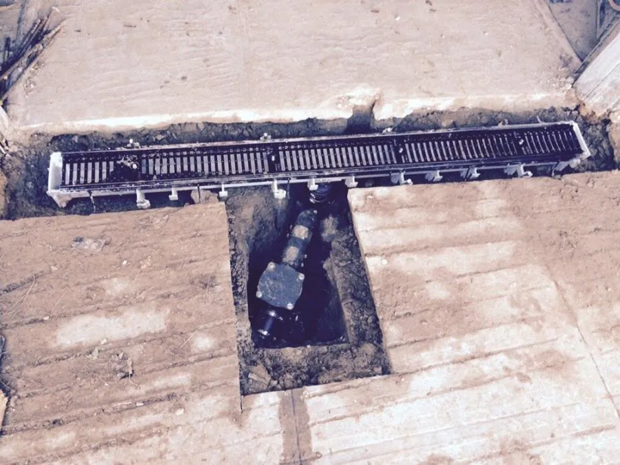 A trench drain is installed in a cut-out section of concrete flooring, with a visible pipe connection beneath.