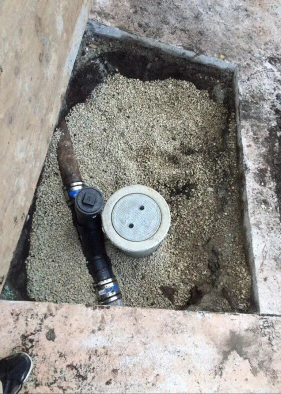 A plumbing cleanout access installed in a gravel bed with a connected pipe, surrounded by concrete surfaces.
