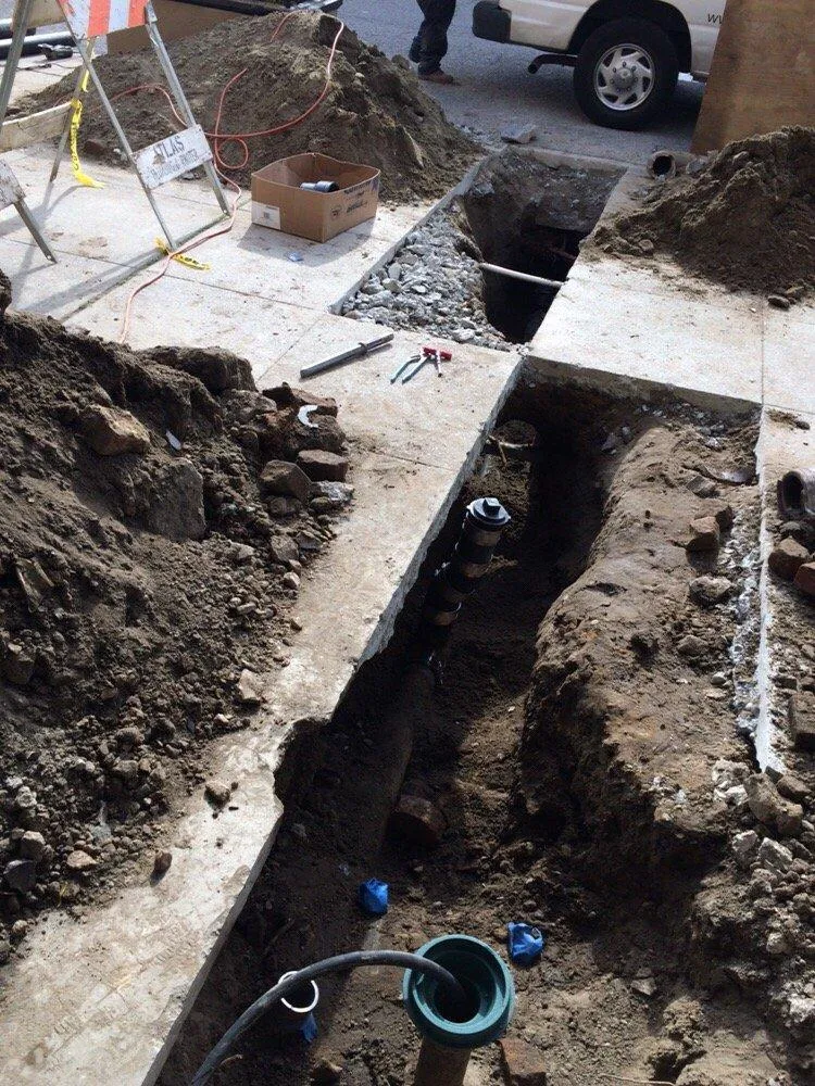  Excavated trench with exposed pipes and tools, surrounded by dirt and concrete. A nearby utility truck and safety cones are visible.