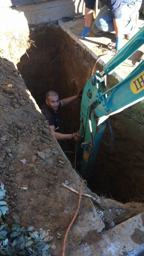  Worker in a deep trench using heavy equipment, with another person above ground. Soil and plants are visible around the trench edges.