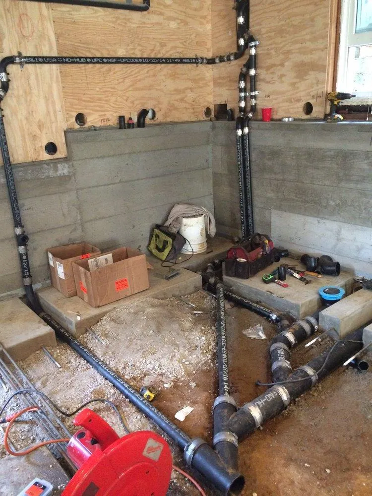  Plumbing installation in progress with black pipes, tools, and materials neatly arranged in a partially constructed room with plywood walls and concrete base.