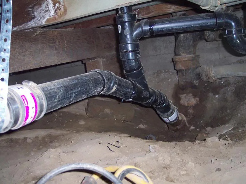  Close-up view of plumbing pipes installed in an underground space with dirt floor and wooden beams.