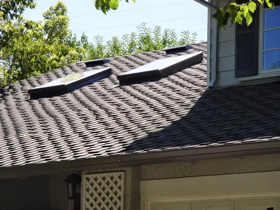 Roof with dark shingles and three skylights under sunlight; part of house exterior visible with window and light.