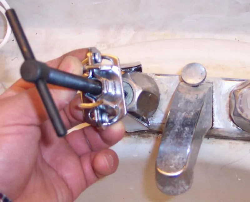 Close-up of a hand using a T-handle wrench on a metal faucet in a bathroom setting.
