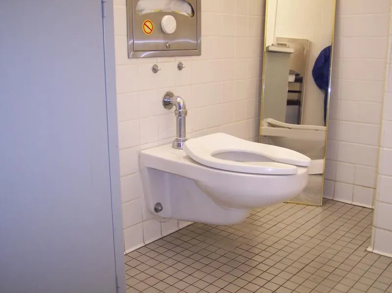  Wall-mounted toilet with visible plumbing against tiled wall and floor. Mirror and partial view of a person in the background.