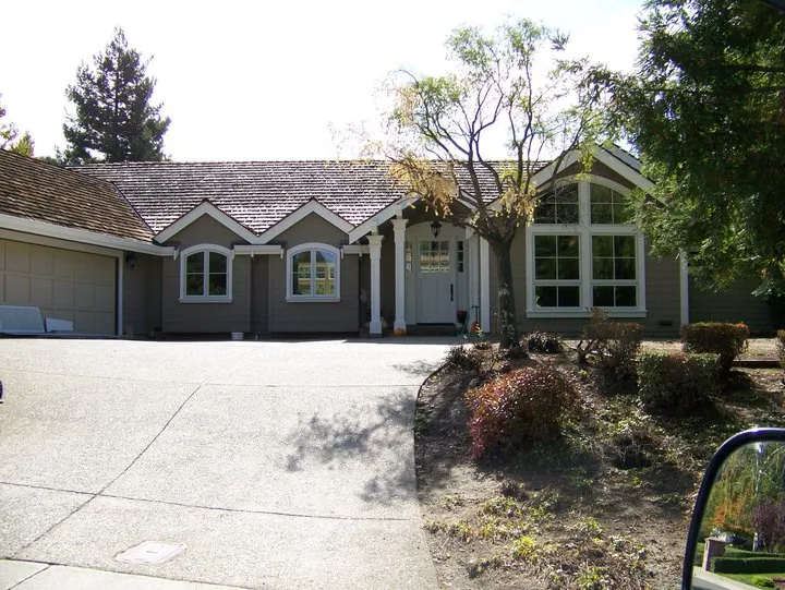 A house with gray siding and a shingled roof, featuring large windows and an arched entryway. Surrounded by trees and shrubs.
