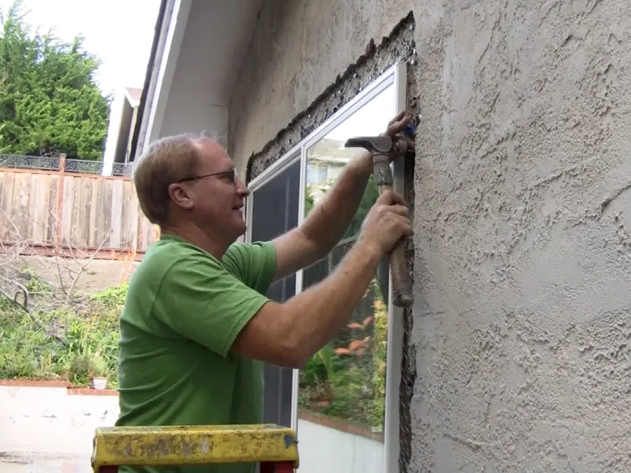 Man in a green shirt uses a hammer to install a window frame on a stucco wall, standing on a yellow ladder. Man in a green shirt uses a hammer to install a window frame on a stucco wall, standing on a yellow ladder.