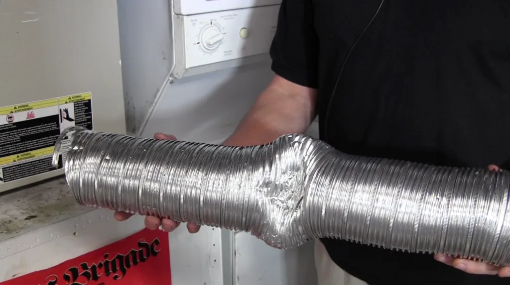 A person holds a bent flexible duct near a dryer, inspecting it for damage.