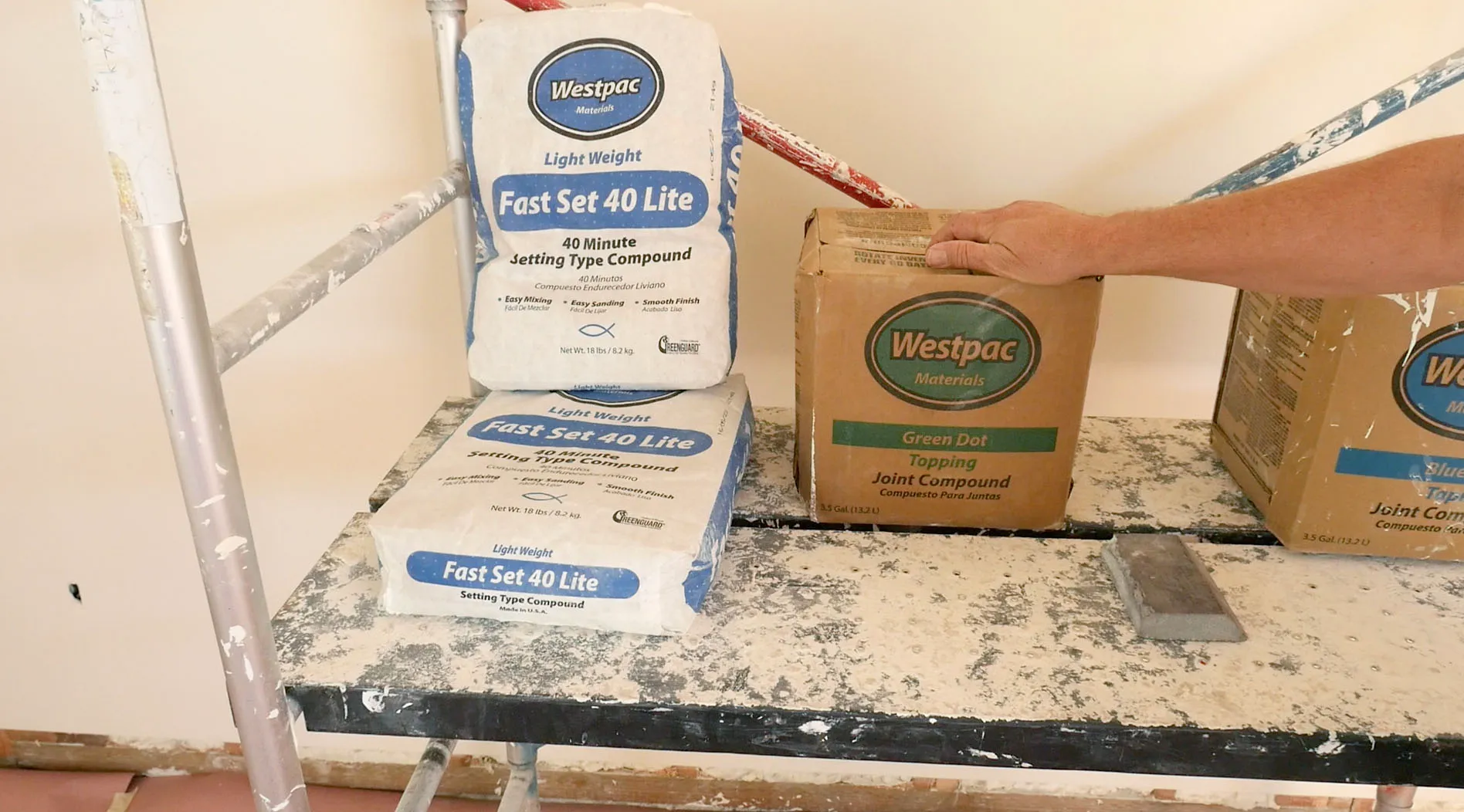  Bags and boxes of joint compound on a paint-splattered scaffold, with a hand steadying a box.