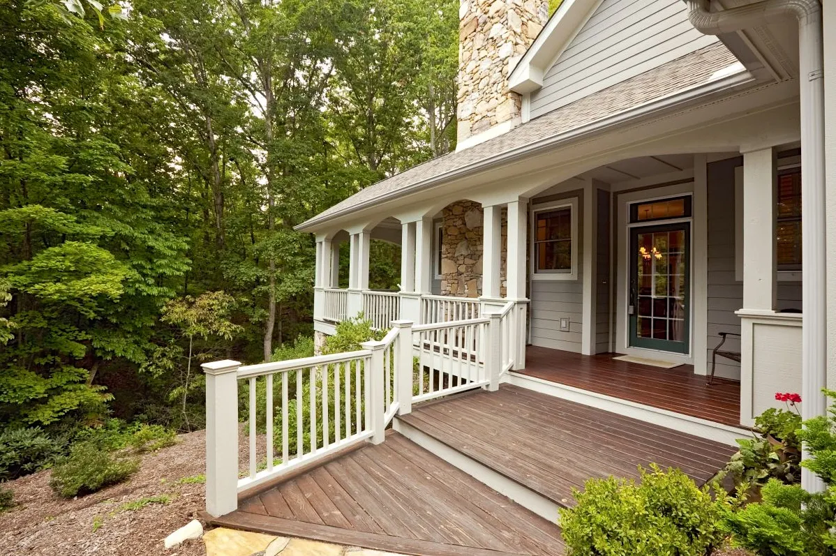  A wooden porch with white railings and a stone exterior wall, set amidst dense green trees.