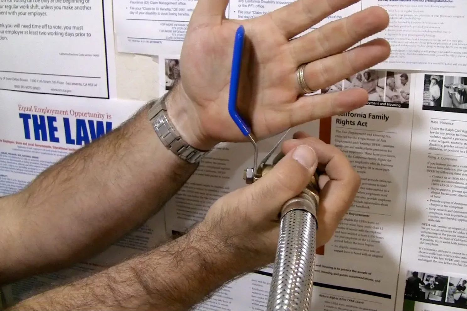 Close-up of hands adjusting a blue-handled valve on a flexible metal hose against a wall with posters. Close-up of hands adjusting a blue-handled valve on a flexible metal hose against a wall with posters.