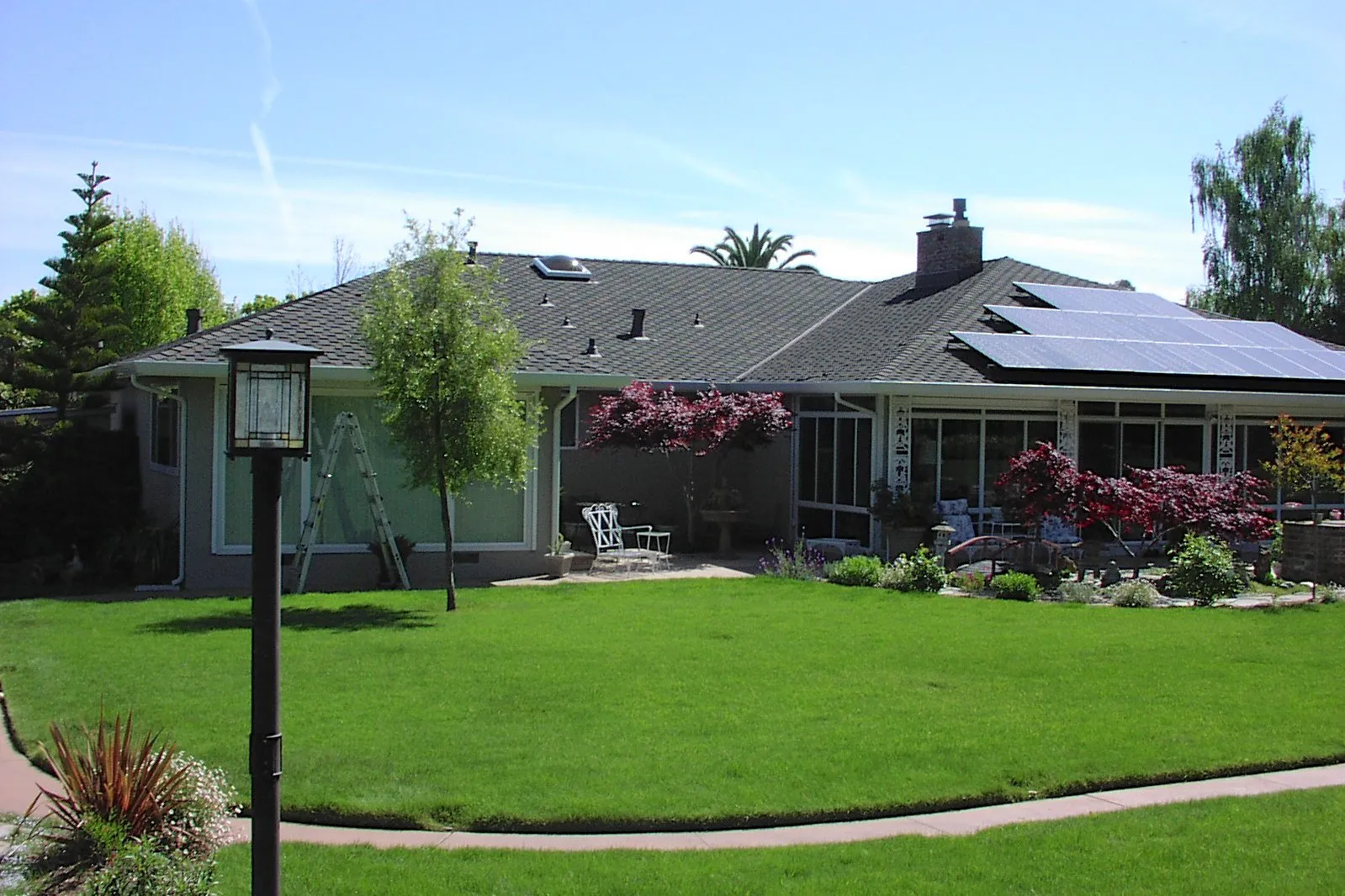 A residential house with solar panels on the roof, a ladder against the wall, green lawn, trees, and garden furniture. A residential house with solar panels on the roof, a ladder against the wall, green lawn, trees, and garden furniture.