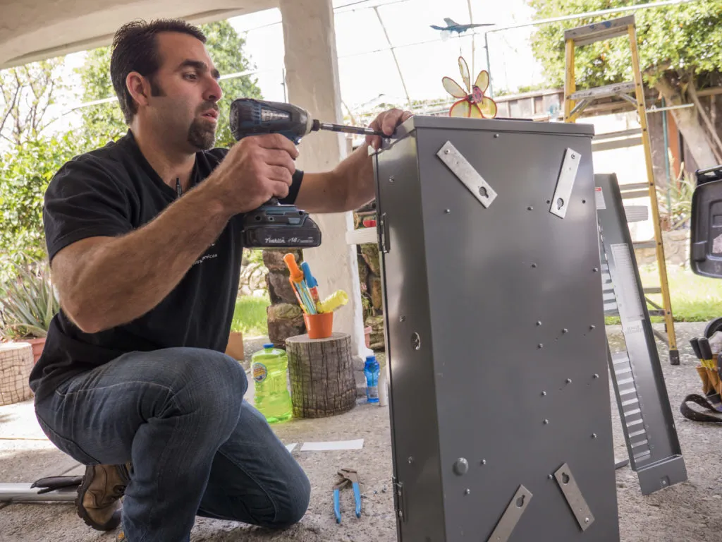 A technician kneels, using a cordless drill on a metal enclosure. Nearby, tools and a ladder are visible, with a garden setting in the background. A technician kneels, using a cordless drill on a metal enclosure. Nearby, tools and a ladder are visible, with a garden setting in the background.