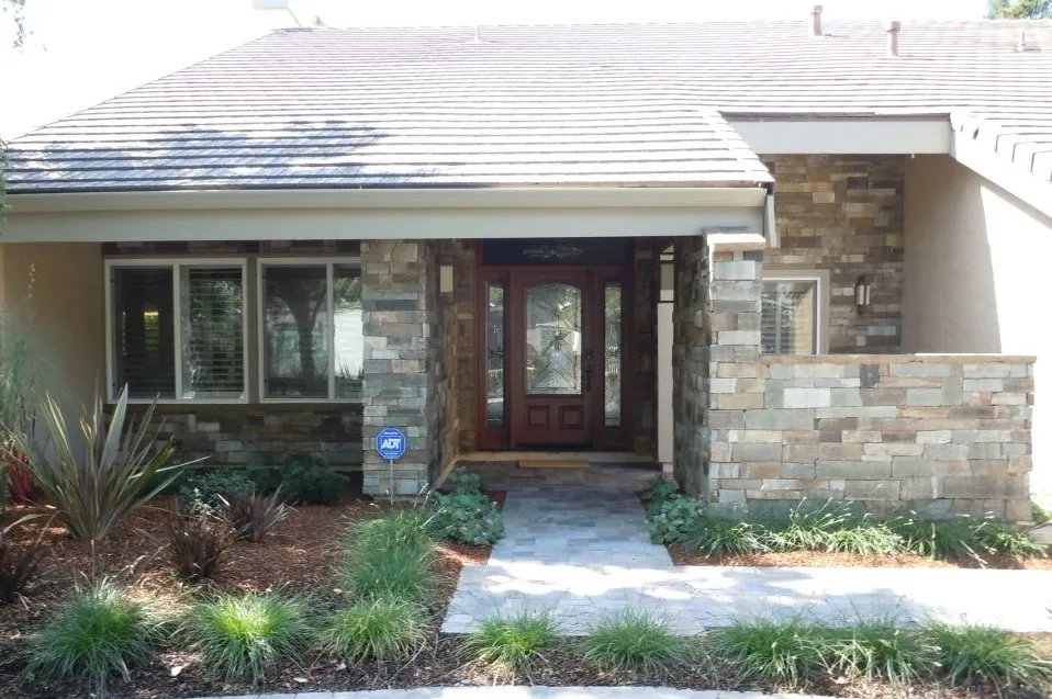  Front view of a house with stone exterior, wooden door with glass panels, surrounded by plants and a small pathway.