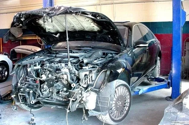  A car with its front bumper and hood removed is elevated on a lift in a garage, revealing the engine and inner workings.