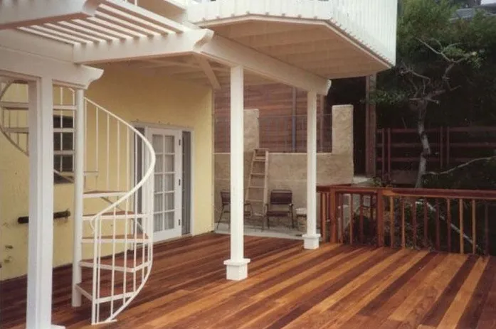 A wooden deck with a spiral staircase, white columns, and a yellow wall with a glass door. A ladder and chairs are in the background. A wooden deck with a spiral staircase, white columns, and a yellow wall with a glass door. A ladder and chairs are in the background.