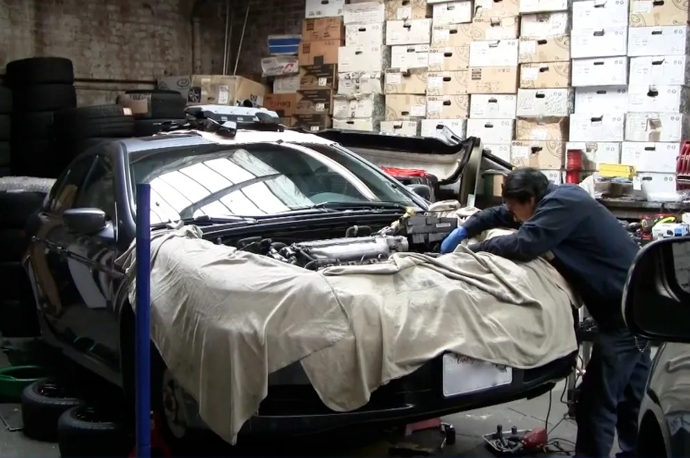 A technician works on a car engine covered with a cloth in a garage, surrounded by stacked boxes and tires. A technician works on a car engine covered with a cloth in a garage, surrounded by stacked boxes and tires.