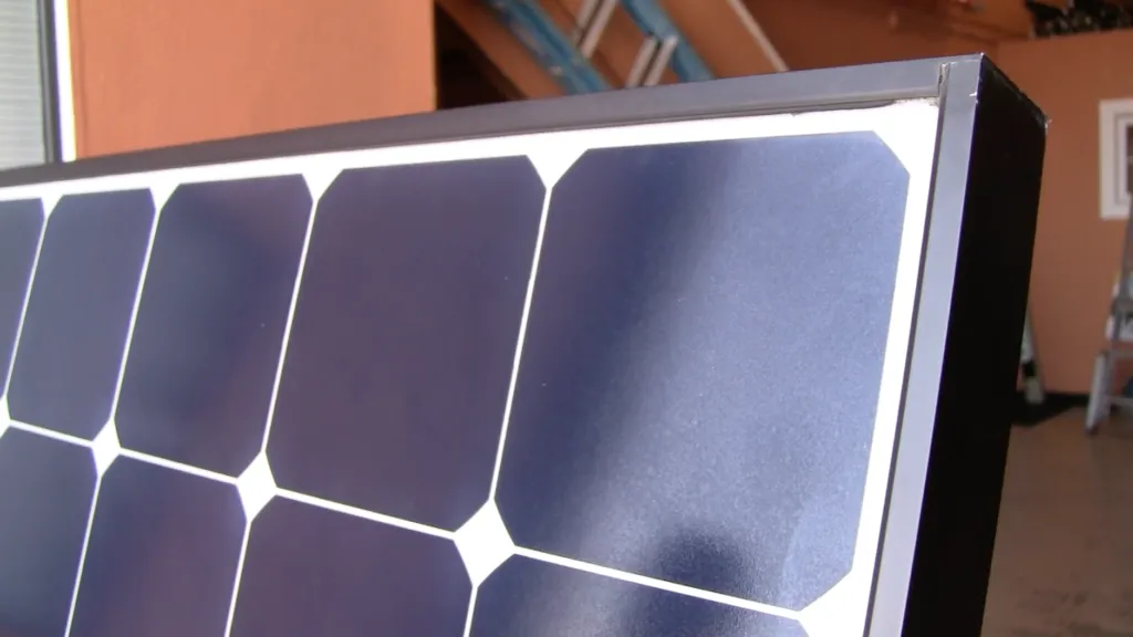 Close-up of a solar panel with black cells and a metal frame, set against an indoor backdrop with ladders and a light-colored wall in the background.