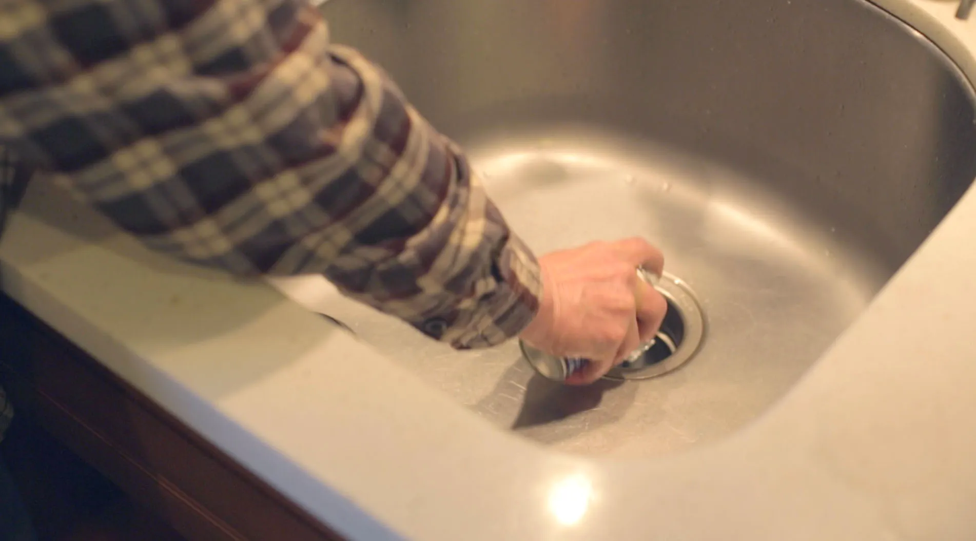 A person in a plaid shirt adjusts a sink stopper in a stainless steel sink. A person in a plaid shirt adjusts a sink stopper in a stainless steel sink.