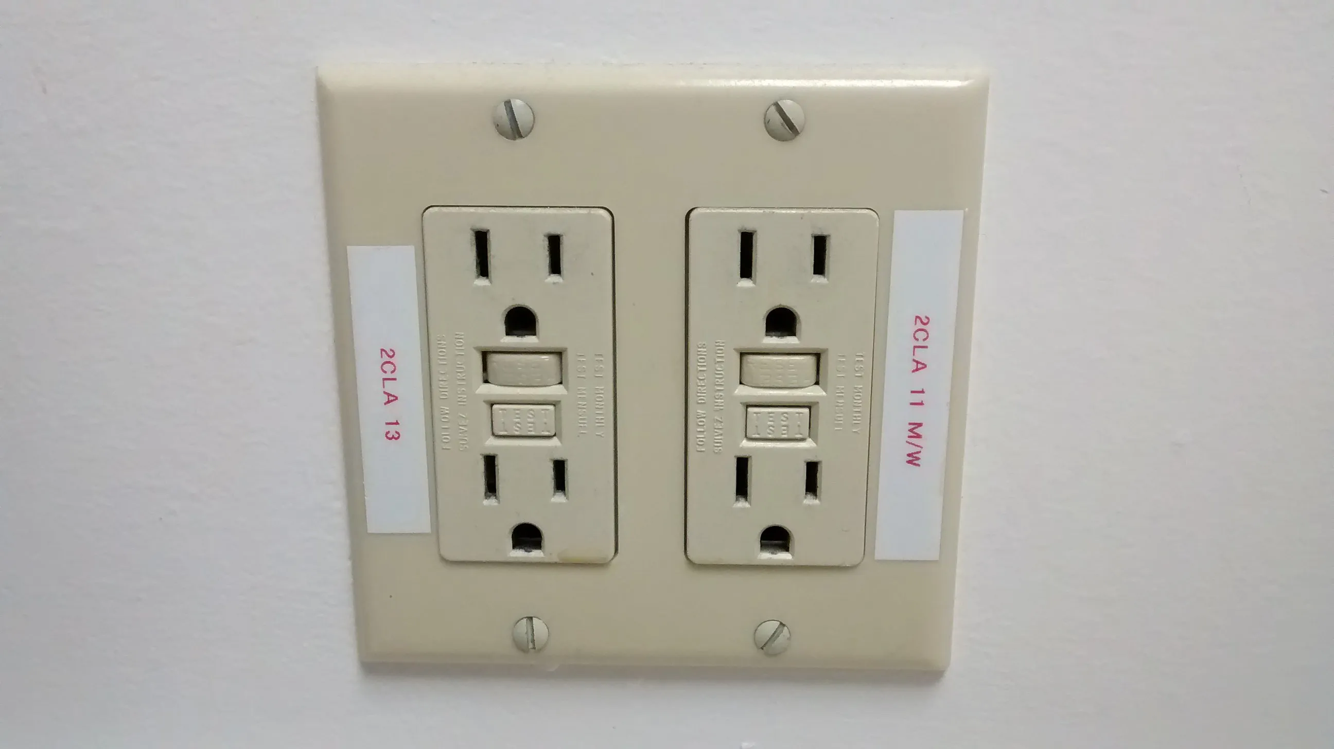 Beige dual electrical outlet with reset buttons and labeled slots on a white wall.