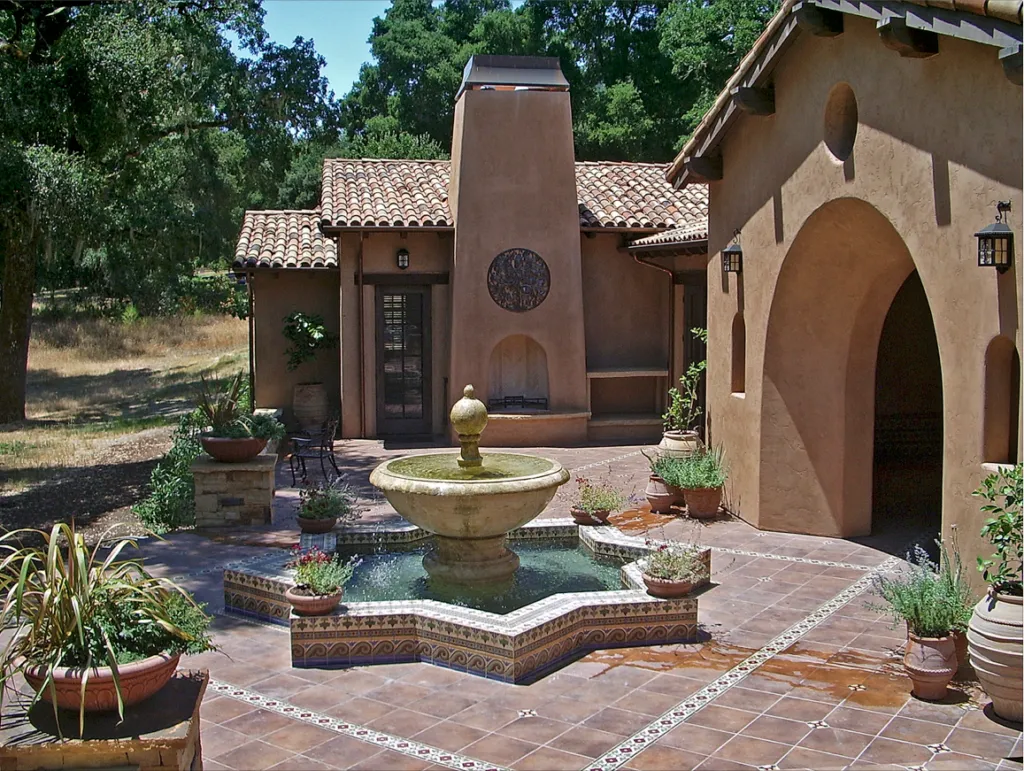 Courtyard with a central fountain, decorative tiles, and potted plants, surrounded by stucco walls and tile roofing. Courtyard with a central fountain, decorative tiles, and potted plants, surrounded by stucco walls and tile roofing.