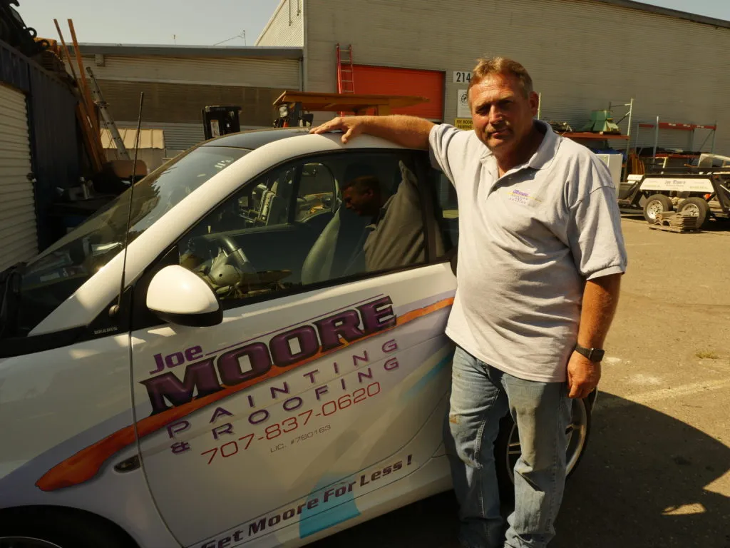  Person stands beside Joe Moore Painting & Roofing vehicle, hand resting on the roof, in an outdoor setting with various equipment in the background.