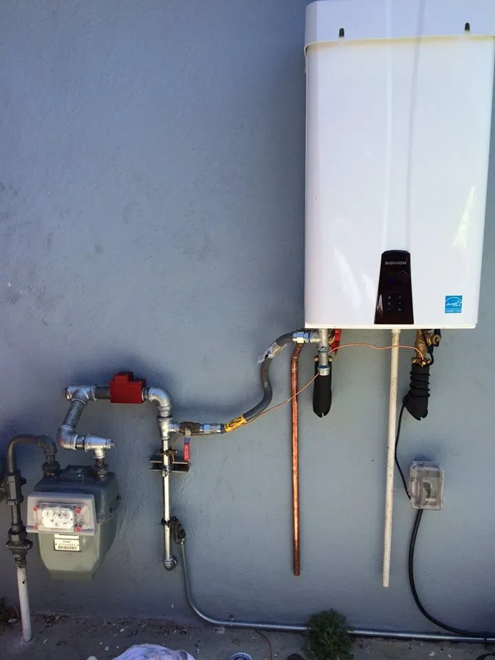 Wall-mounted tankless water heater with connected pipes and a nearby gas meter against a blue wall.