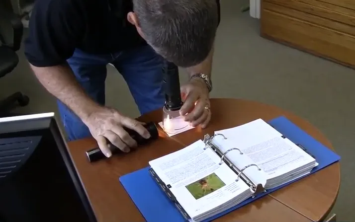 A person inspects a sample using a magnifying tool over a table with an open binder and book. A person inspects a sample using a magnifying tool over a table with an open binder and book.