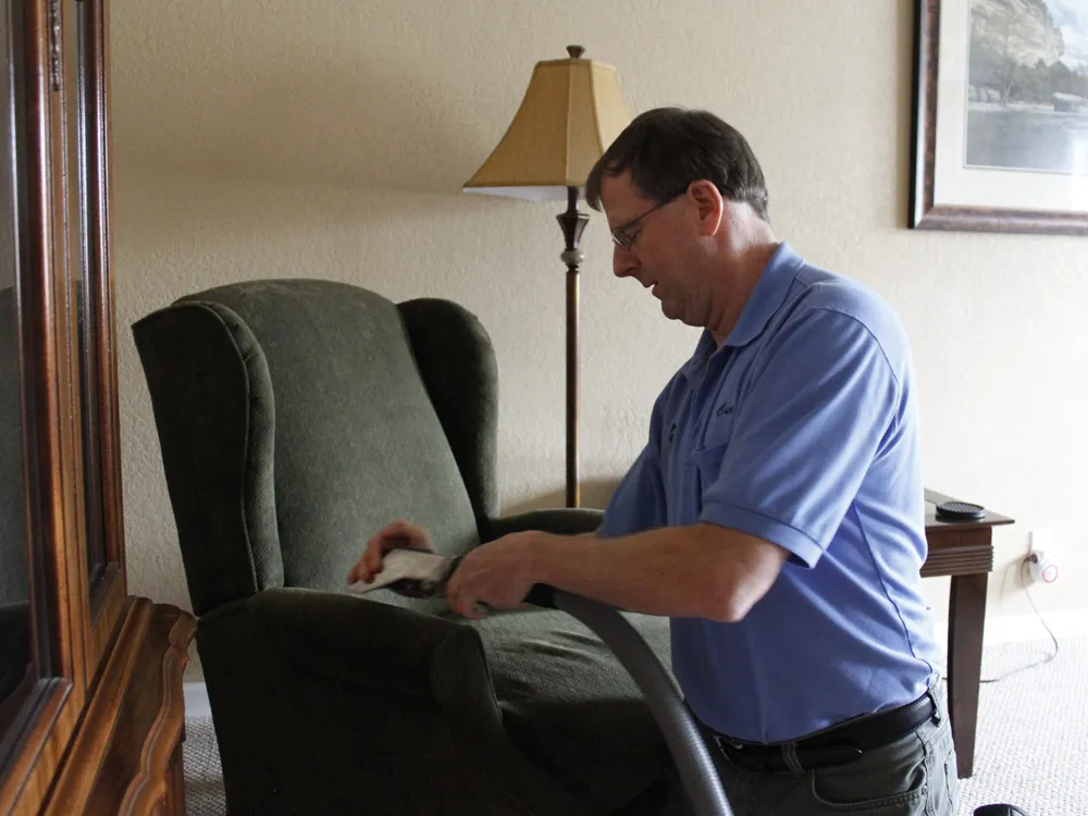 A person in a blue shirt uses a vacuum to clean a green armchair in a room with a lamp and artwork. A person in a blue shirt uses a vacuum to clean a green armchair in a room with a lamp and artwork.