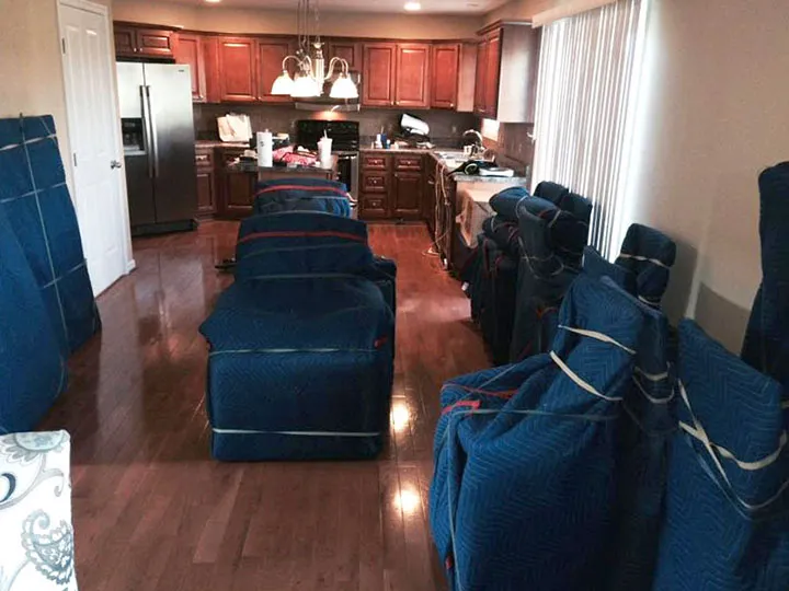 Furniture wrapped in blue moving blankets arranged neatly in a kitchen with wooden cabinets and appliances. Furniture wrapped in blue moving blankets arranged neatly in a kitchen with wooden cabinets and appliances.