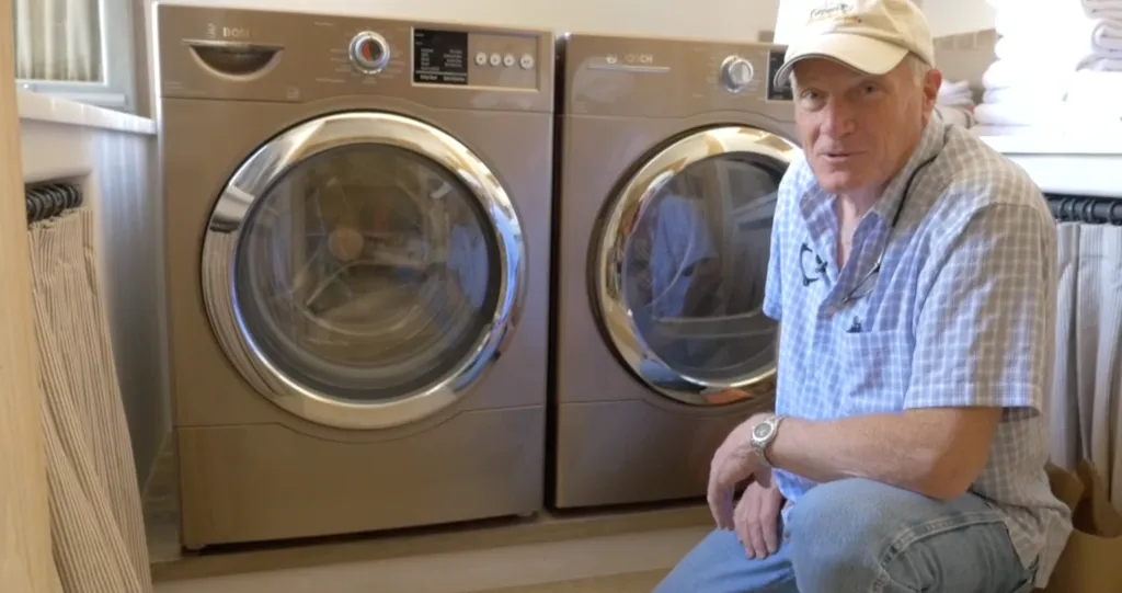 A person kneels next to a pair of front-loading Bosch laundry machines in a well-organized room with folded towels nearby. A person kneels next to a pair of front-loading Bosch laundry machines in a well-organized room with folded towels nearby.
