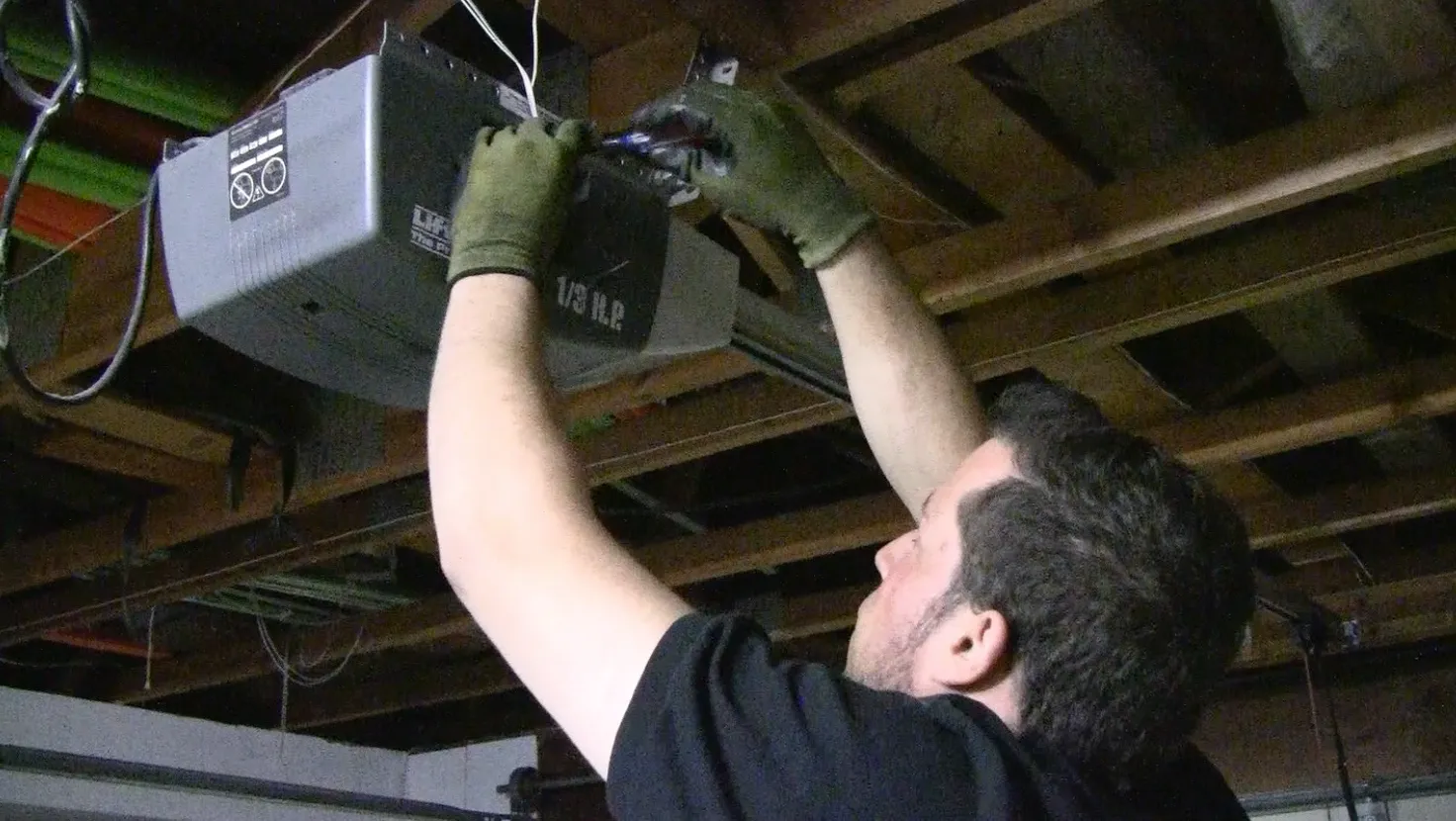 A technician in gloves works on a garage door opener mounted to a ceiling with exposed wooden beams. Tools are in use. A technician in gloves works on a garage door opener mounted to a ceiling with exposed wooden beams. Tools are in use.