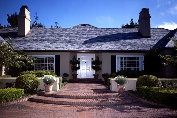A well-maintained house with a slate roof, two chimneys, large windows, and a brick walkway leading to a white front door, surrounded by potted plants and manicured hedges.