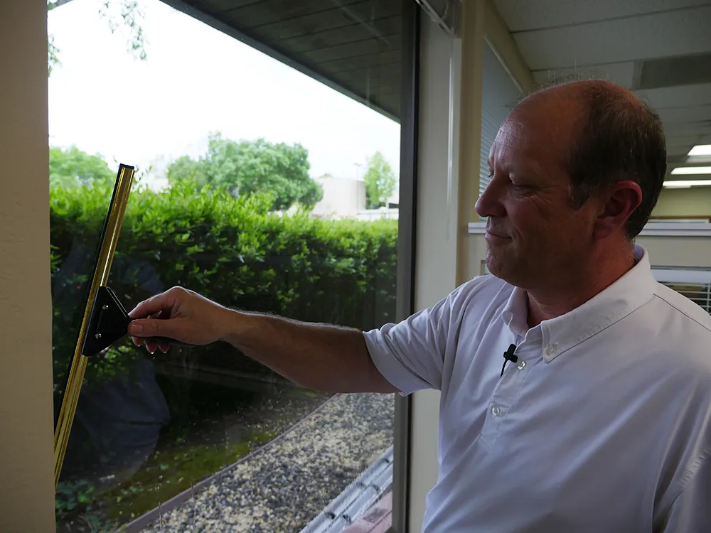 A person in a white shirt uses a squeegee to clean a large window, with greenery visible outside. A person in a white shirt uses a squeegee to clean a large window, with greenery visible outside.