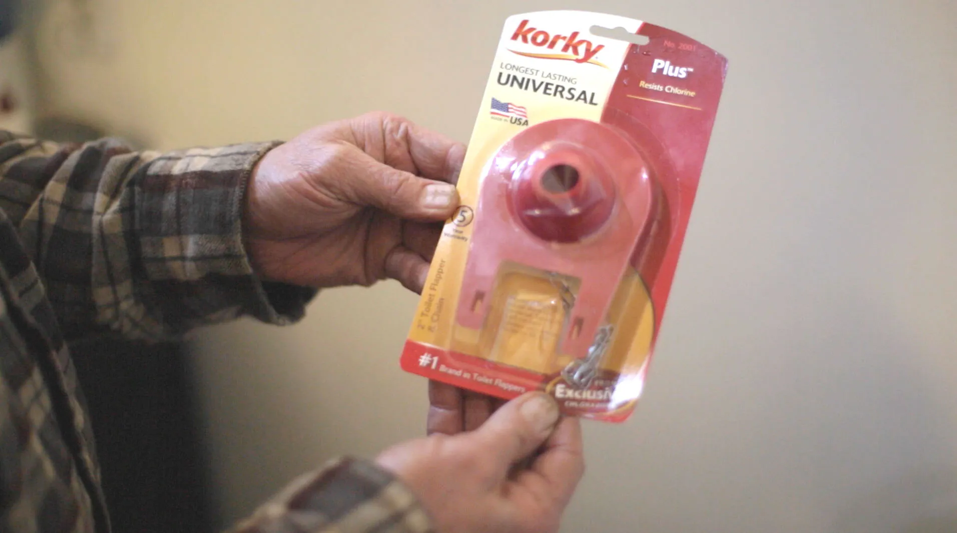Hands holding a packaged Korky universal toilet flapper against a plain background. Hands holding a packaged Korky universal toilet flapper against a plain background.