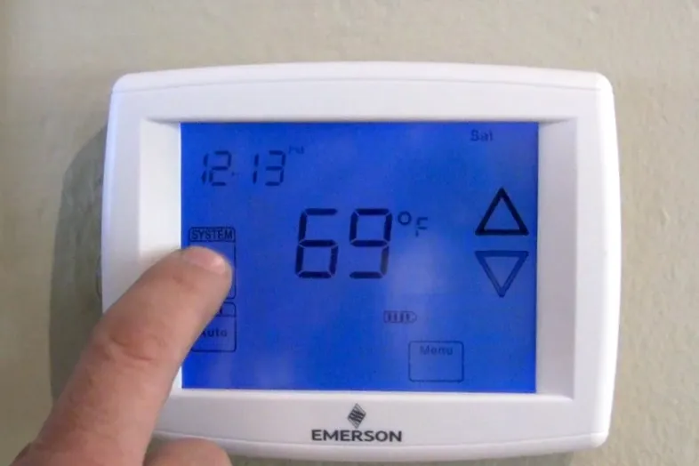 A hand adjusting a digital thermostat showing 69°F with a blue screen and white casing. A hand adjusting a digital thermostat showing 69°F with a blue screen and white casing.