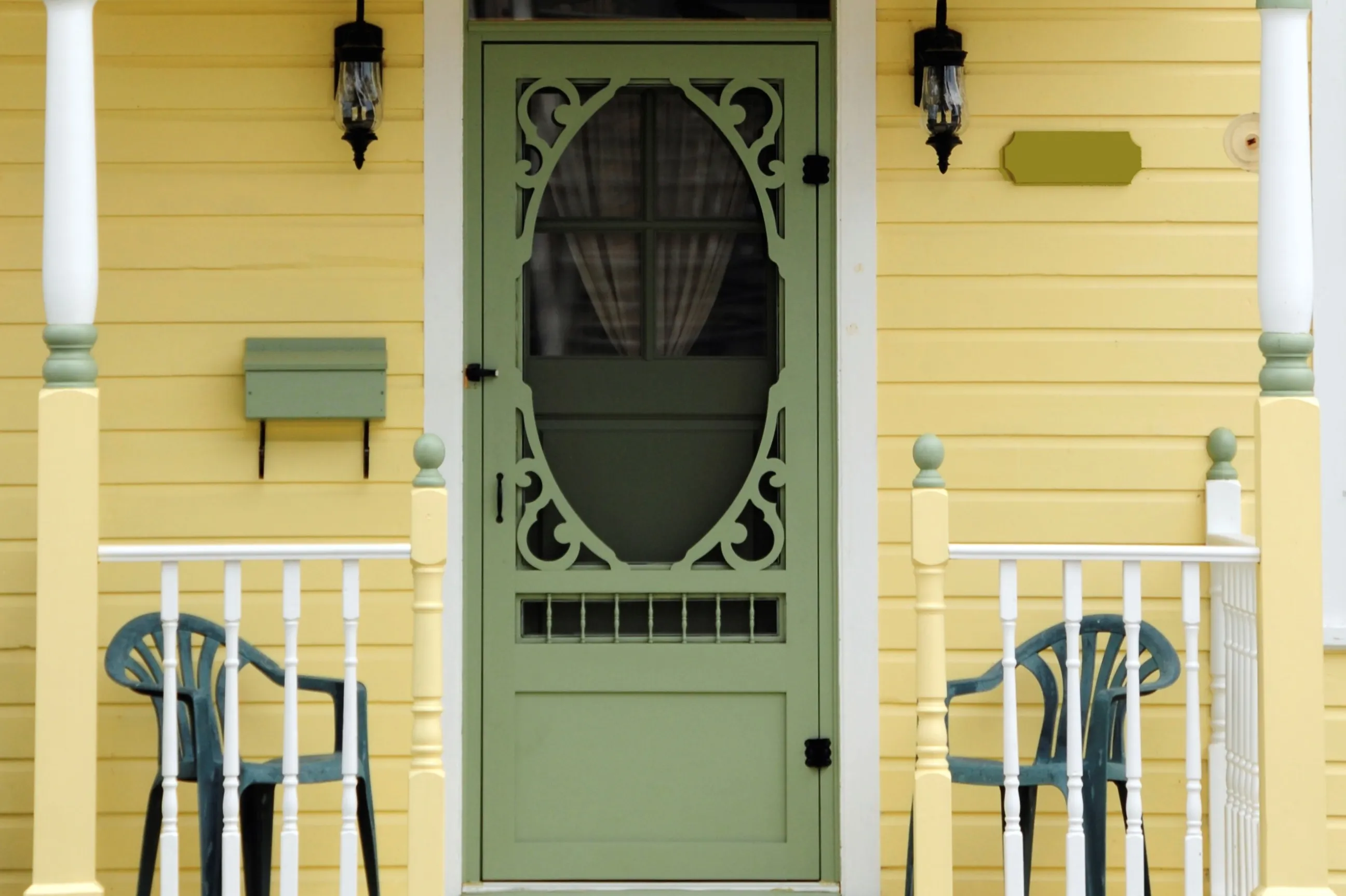 Green screen door with intricate design on yellow house, flanked by two plastic chairs and porch railings. Green screen door with intricate design on yellow house, flanked by two plastic chairs and porch railings.