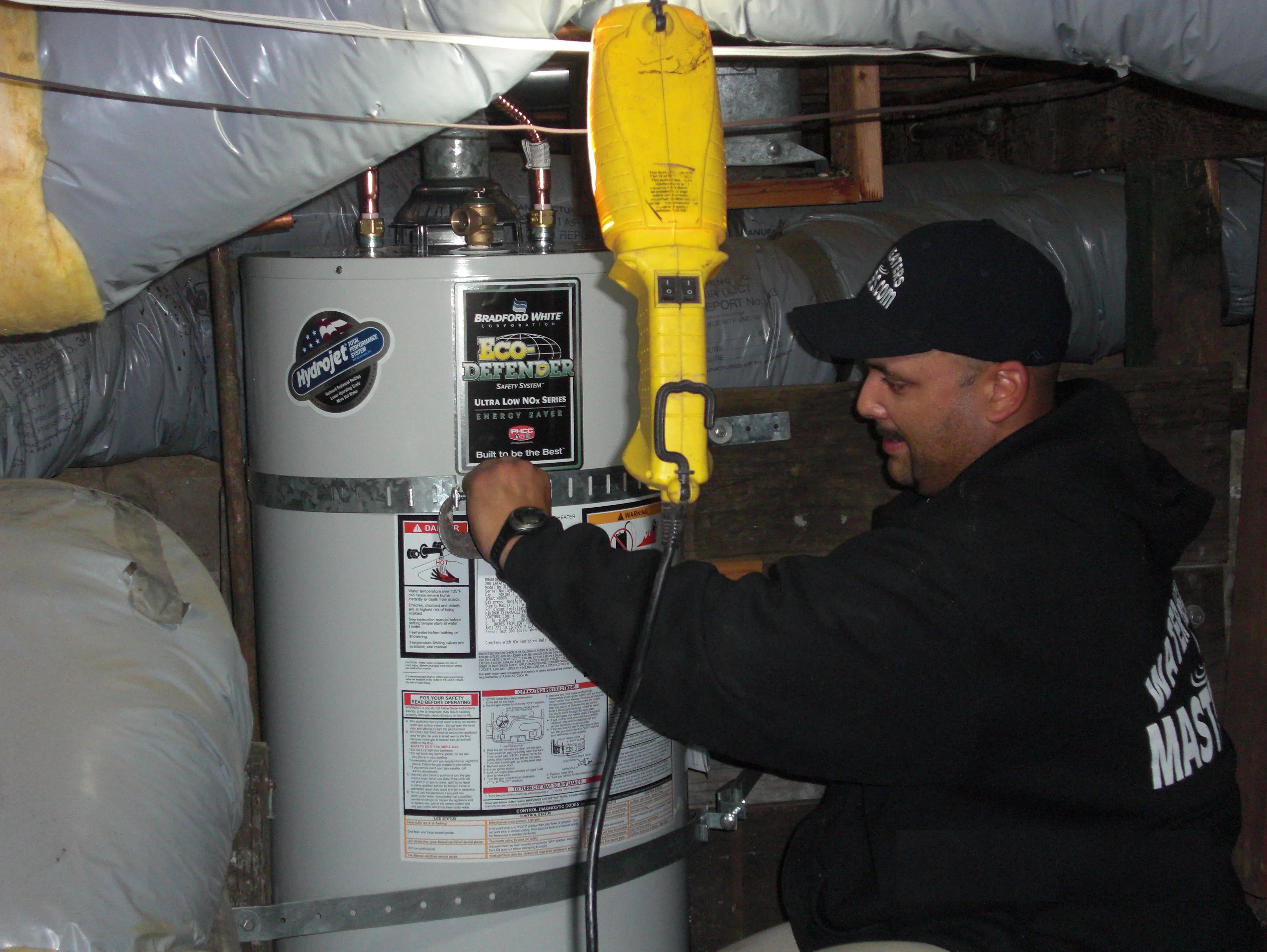 Technician adjusting a water heater in a small space, surrounded by ducts, wearing a Water Heaters Masters Inc. hoodie and cap, using tools. Technician adjusting a water heater in a small space, surrounded by ducts, wearing a Water Heaters Masters Inc. hoodie and cap, using tools.
