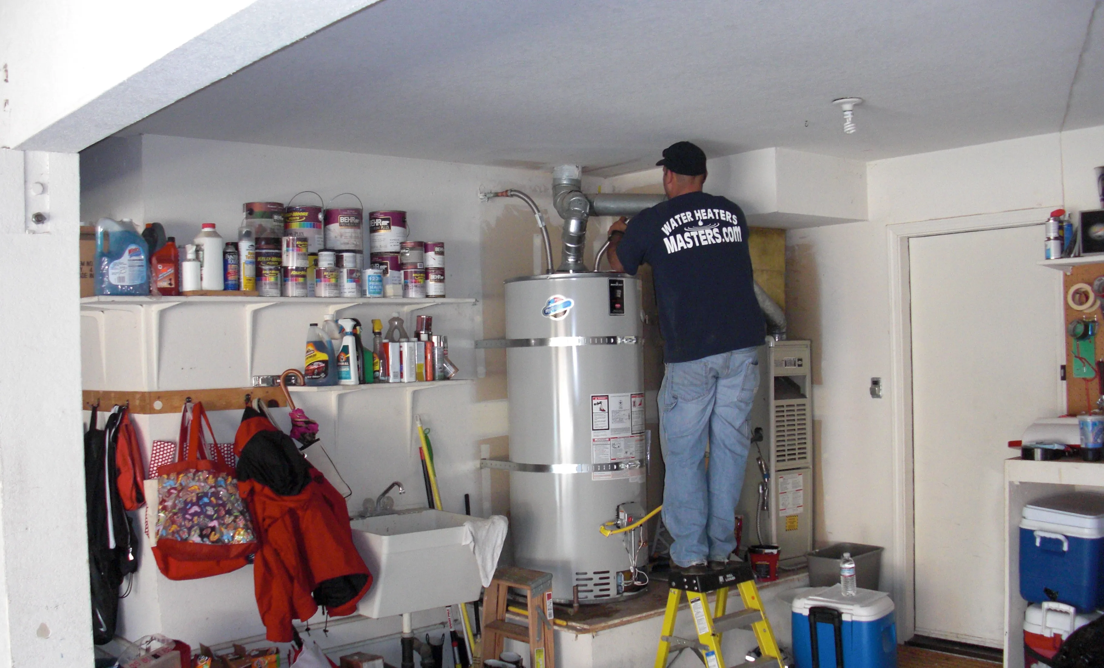 Technician from Water Heaters Masters Inc. stands on a ladder, installing a water heater in a garage filled with shelves, tools, and cleaning supplies. Technician from Water Heaters Masters Inc. stands on a ladder, installing a water heater in a garage filled with shelves, tools, and cleaning supplies.