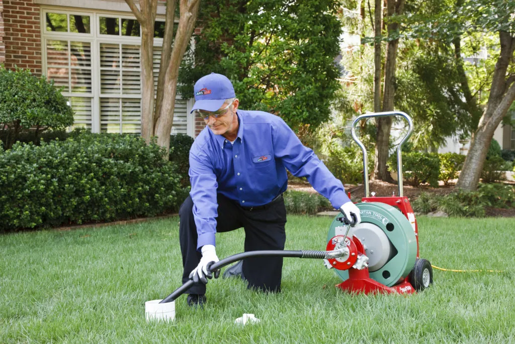  Technician using a drain cleaning machine on a lawn, with a tool beside him. A brick house and trees are in the background.
