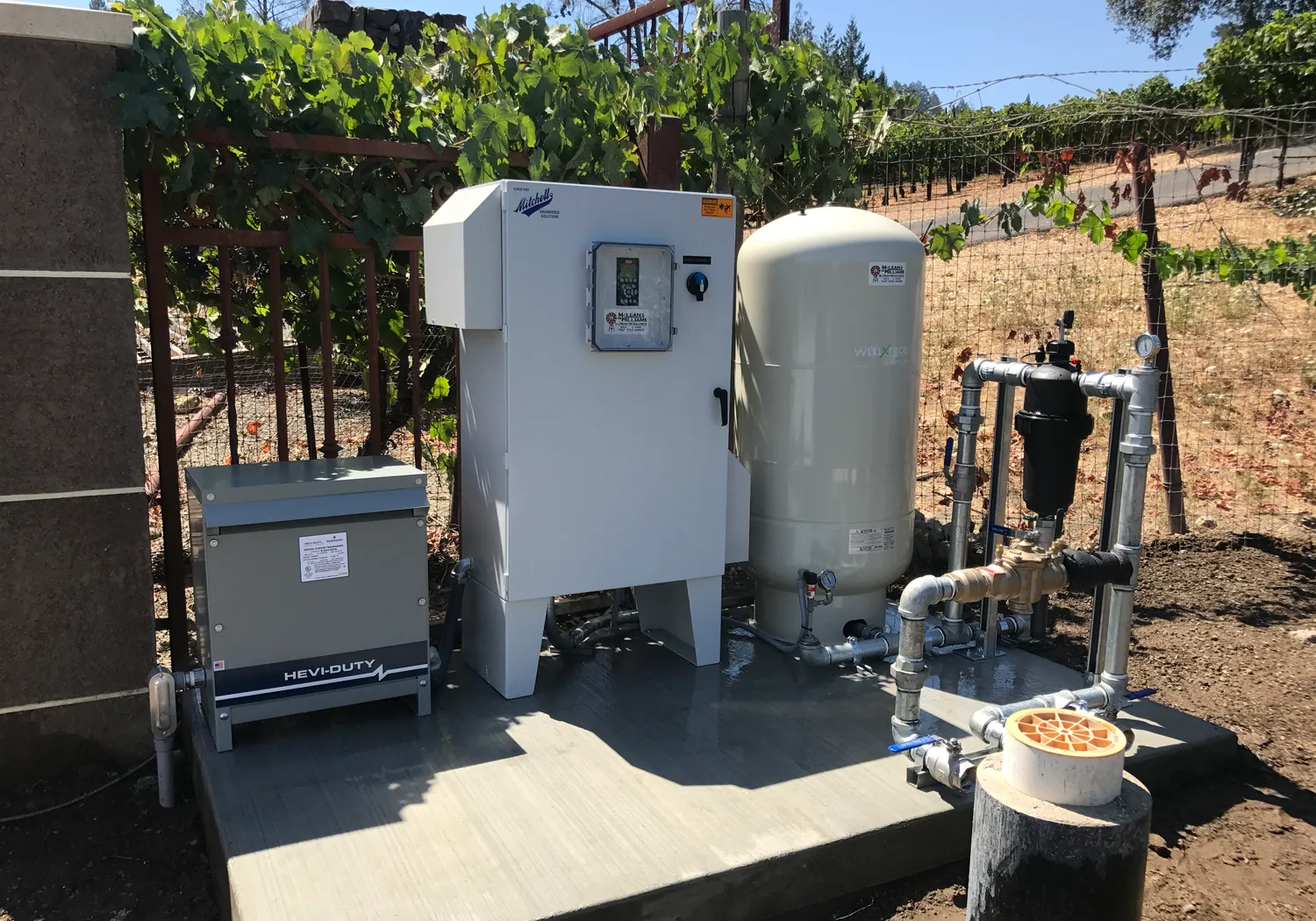 Irrigation system setup with control boxes, pipes, and tank near a vineyard fence. Shadows indicate a sunny day. Irrigation system setup with control boxes, pipes, and tank near a vineyard fence. Shadows indicate a sunny day.