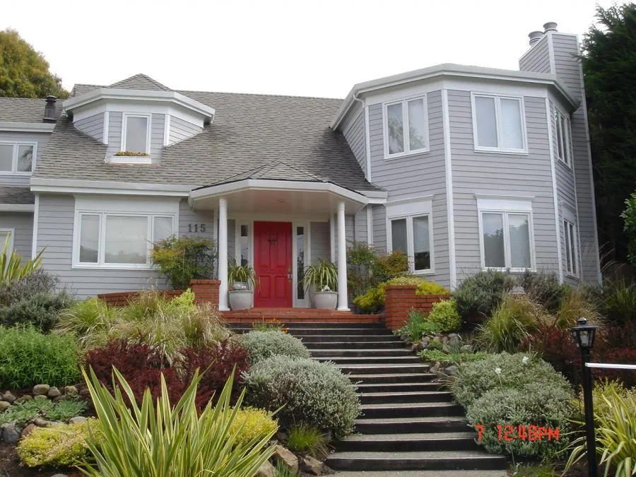  Gray house with a red door, surrounded by well-kept shrubs and plants, featuring a brick staircase leading to the entrance.